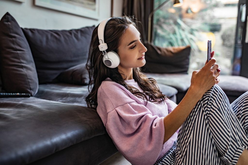 Photo of a young woman sitting on the floor in the living room, using smart phone and listening music