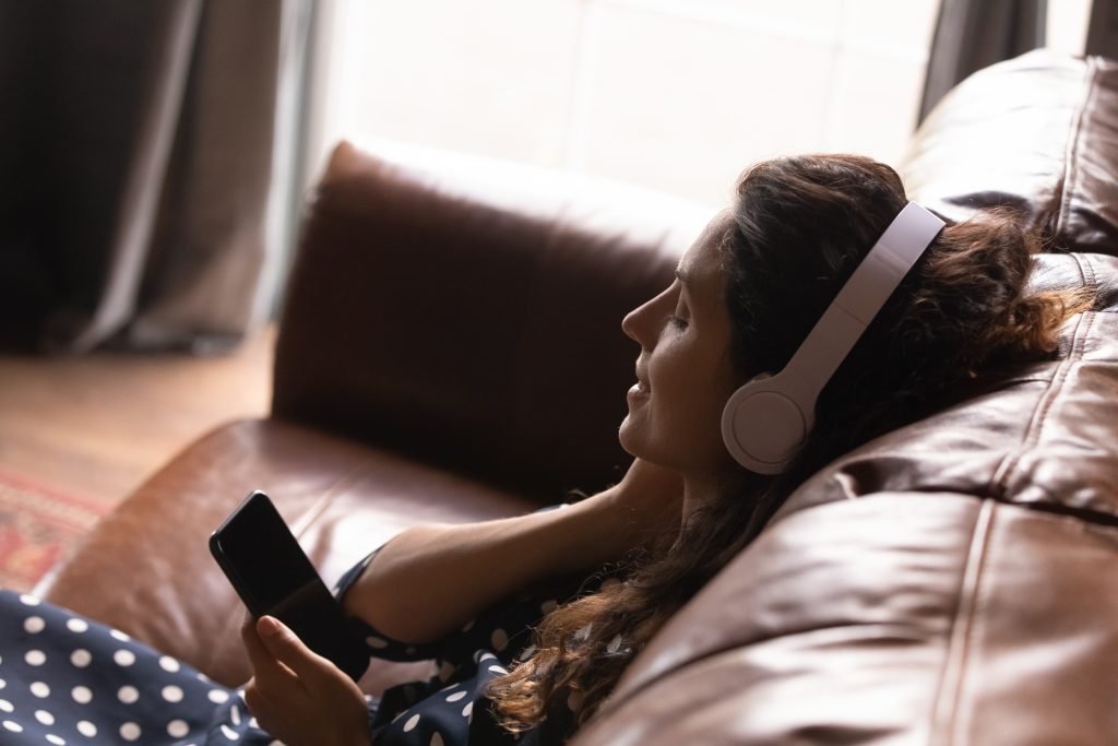 Nesta imagem podemos ver uma moça sentada no sofá escutando um podcast. Nos podcasts e nas rádios onlines é possível veicular mídia display, que é um tipo de mídia programática.