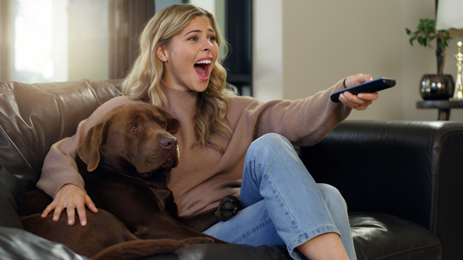 Nesta imagem podemos ver uma moça assistindo TV ao lado de seu cachorro. Ela parece bem animada, enquanto acompanha uma publicidade na TV aberta.
