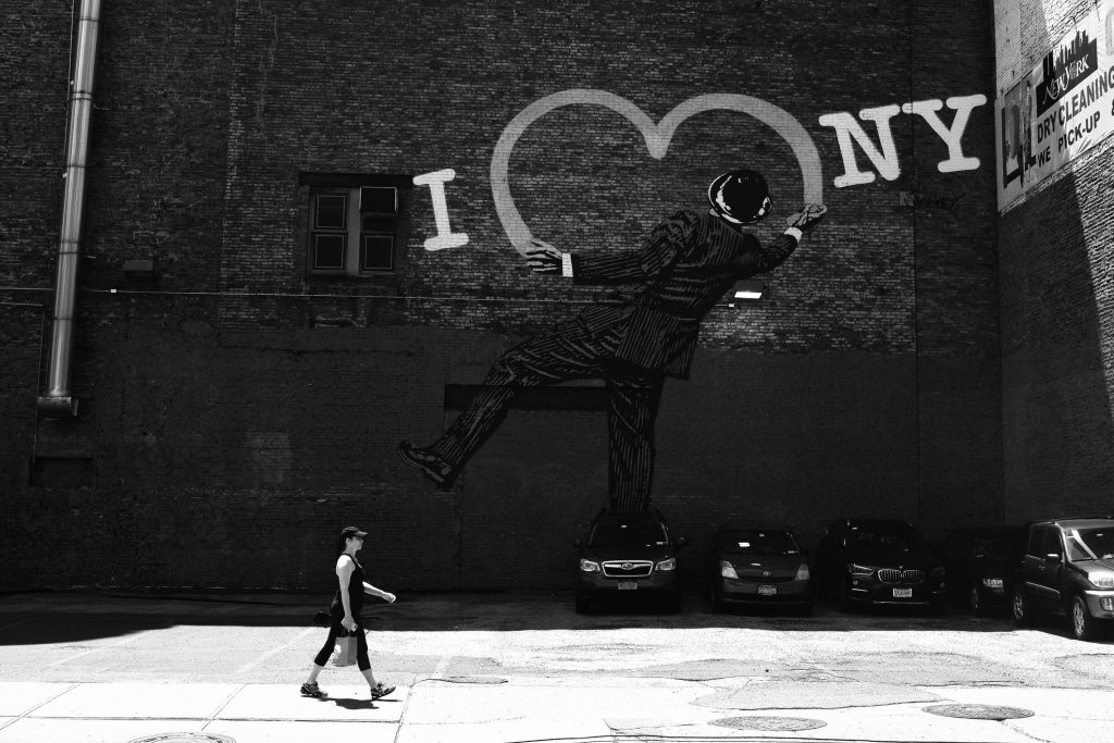 Mulher caminhando na frente de uma parede que está pintada. Na pintura é possível ver um cara pixando o slogan "i love NY", típico da construção do place branding da cidade.