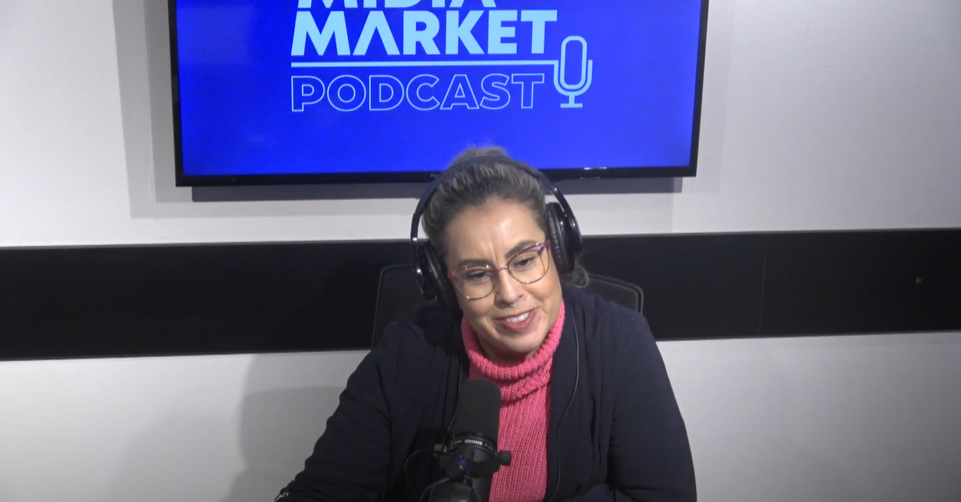 Nesta imagem podemos ver Samanta Tavares, Diretora de Estratégia e Mídia na Exit, no Mídia Market Podcast falando sobre Brandformance. No cenário temos uma televisão com a logo da Mídia Market em azul e branco. Samanta está de óculos e com uma blusa rosa de gola alta.
