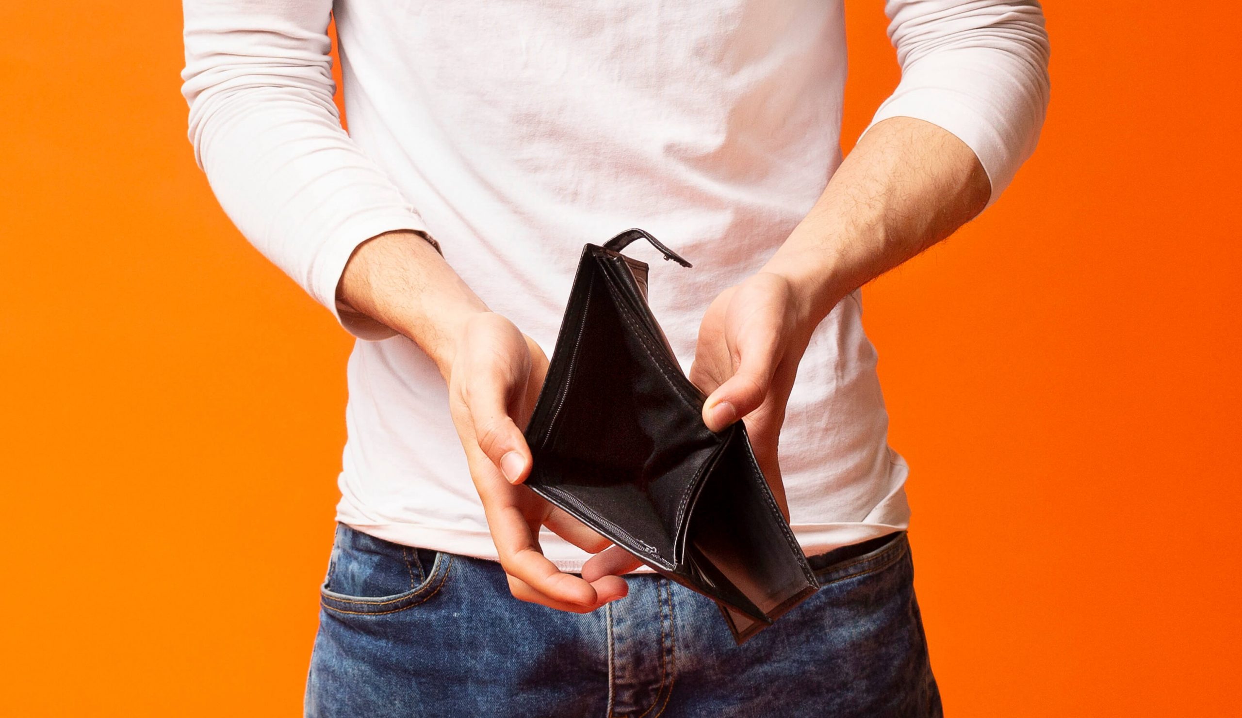 Aqui vemos um homem segurando uma carteira sem dinheiro dentro. Ele está usando calça jeans, camisa branca e o fundo da foto é laranja