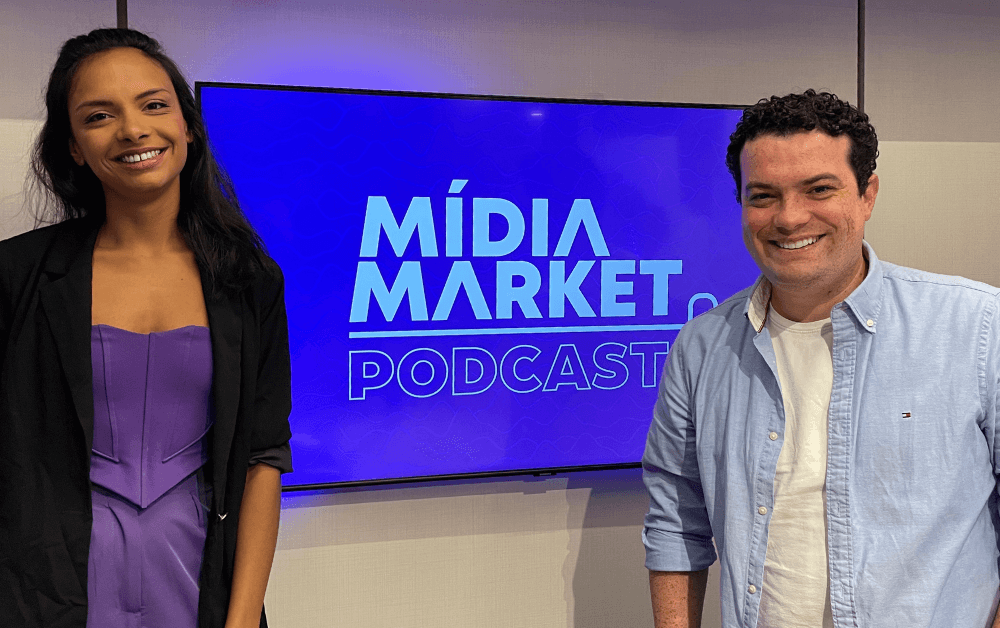 Luiza Telexa com vestido roxo e Otávio pinto com camiseta branca e camisa azul posam para foto em frente a uma televisão com a logo do mídia market podcast de fundo