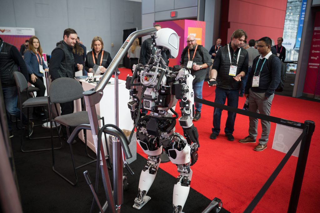 Um grupo de seis pessoas, entre elas, homens e mulheres, conversa entre si enquanto estão reunidas em pé ao redor de um robô.