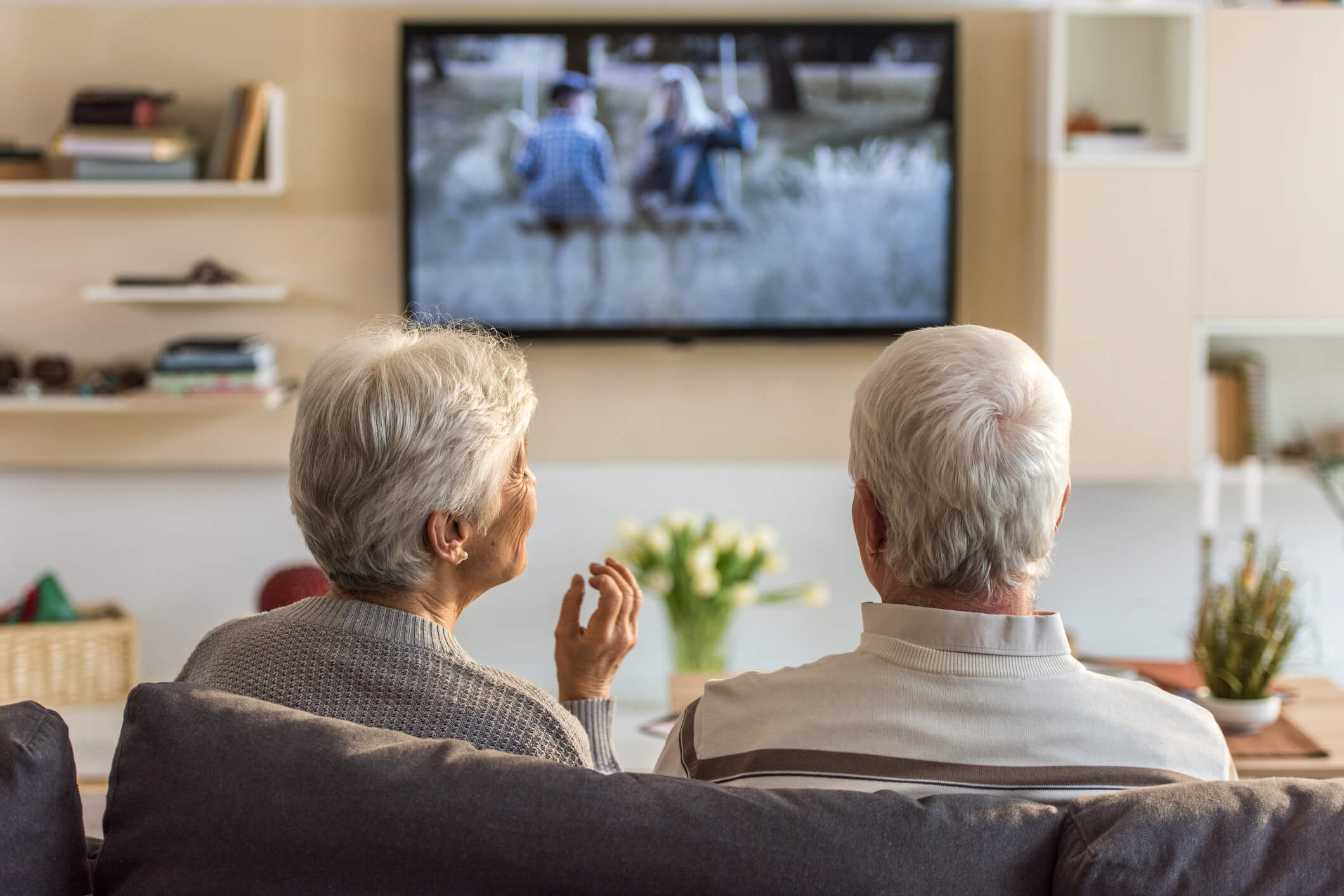 Duas pessoas mais velhas, um homem e uma mulher sentados de costas no sofá assistindo televisão, falando sobre o tipo de mídia televisiva