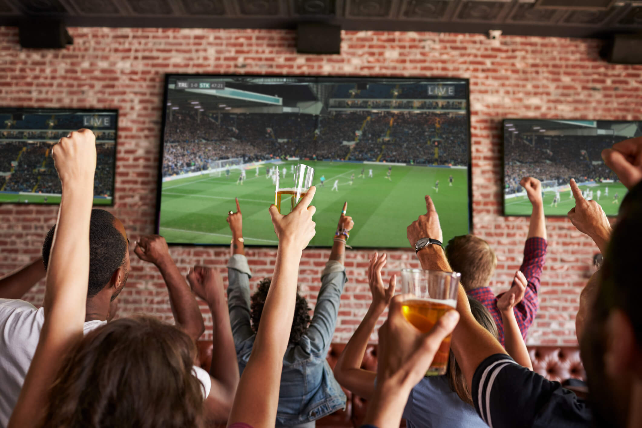 Pessoas assistindo jogos de esporte na Tv e conversando sobre mídia televisiva. Na imagem temos uma TV grande e uma parede 