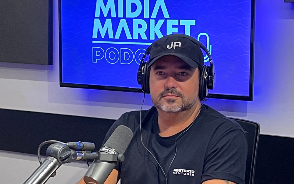Juan Pablo sentado em uma cadeira usando um boné preto e camiseta preta, na imagem temos um microfone e uma TV com a logo do mídia market podcast