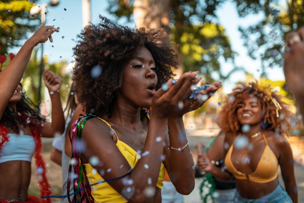 Uma mulher negra com blusa de alça amarela assopra confetes que estão alocados na palma de suas mãos. Ela está em um bloquinho de rua no Carnaval e atrás de si é possível ver alguns foliões desfocados.