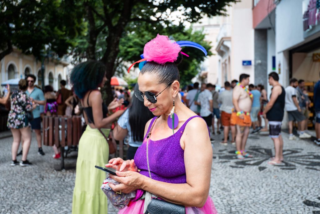 Uma mulher de meia idade está vestindo um body roxo e está com um arranjo de flor rosa no cabelo.Ela está mexendo no celular e está no Carnaval. As redes sociais são ótimos lugares para expor uma campanha de Carnaval.
