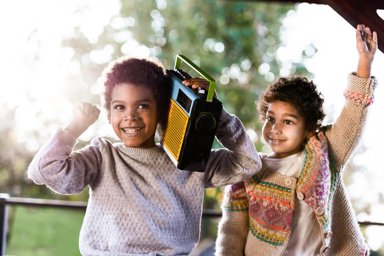 na imagem vemos duas Crianças ouvindo rádio no brasil, uma segura o rádio no ombro, as duas estão em um cenário com árvores
