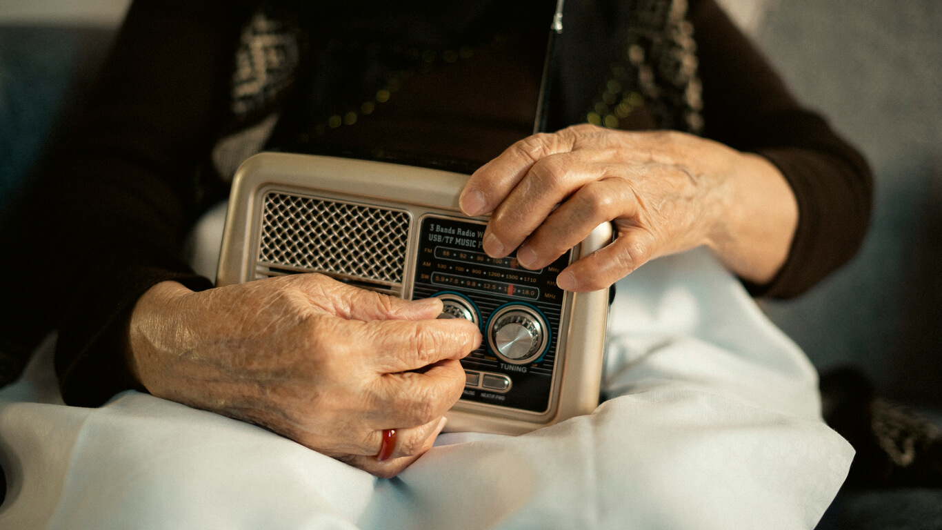 Pessoa idosa sentada com um rádio no colo. Só conseguimos ver as mãos e o rádio. Consumo de rádio no brasil