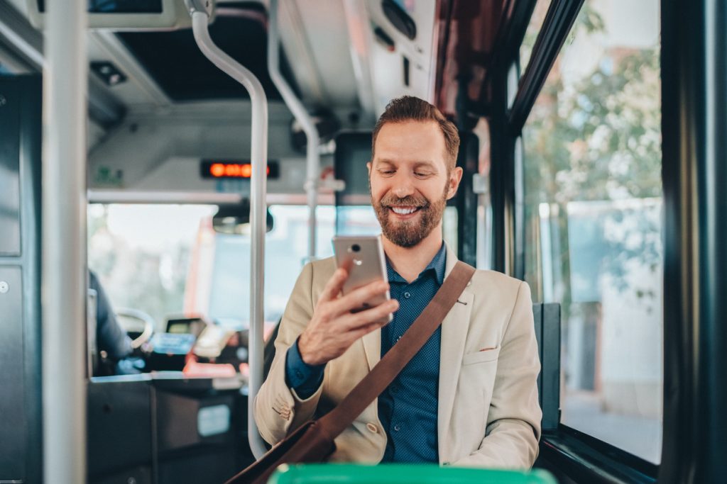 Homem branco e ruivo na casa dos 30 anos está sentado em um banco de ônibus e ao mesmo tempo está mexendo celular.