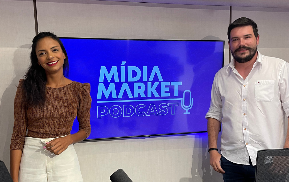Luiza telexa com uma blusa marrom e Pedro pierotti com blusa branca posam para foto ao lado de uma tv com a logo do mídia market podcast