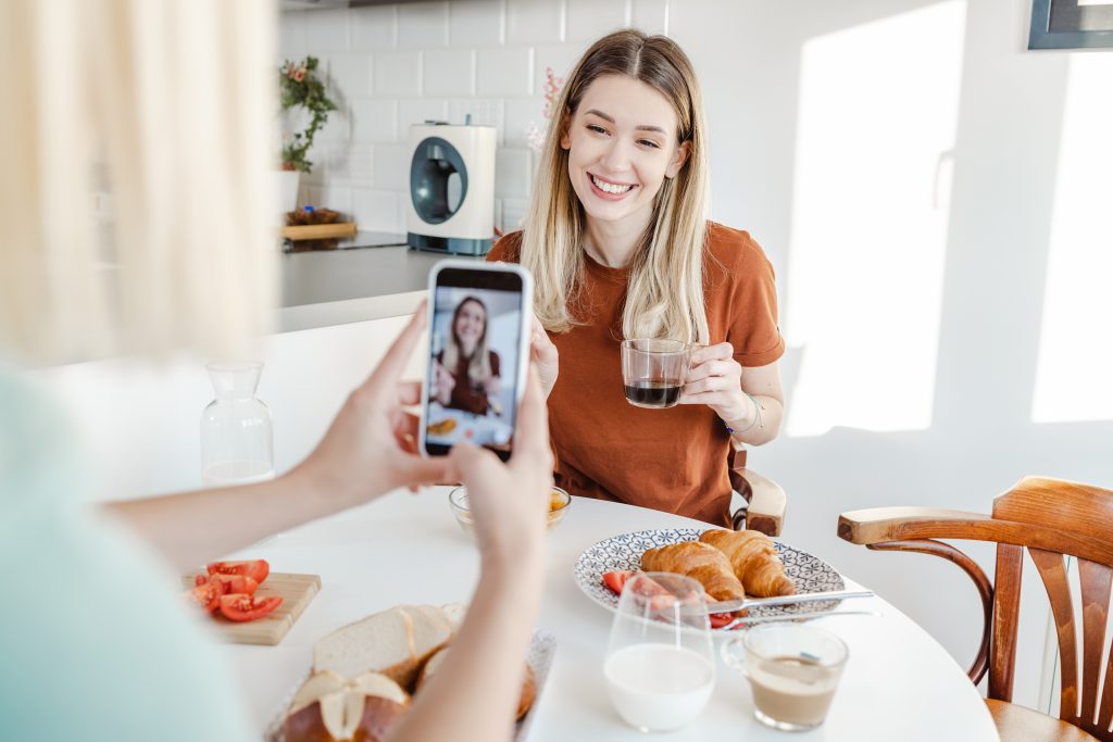Uma mulher branca, loira e jovem está segurando uma xícara de café e sorri para uma foto que está sendo tirado dela através da câmera d eum celular. Ela está preduzindo conteúdo UGC.