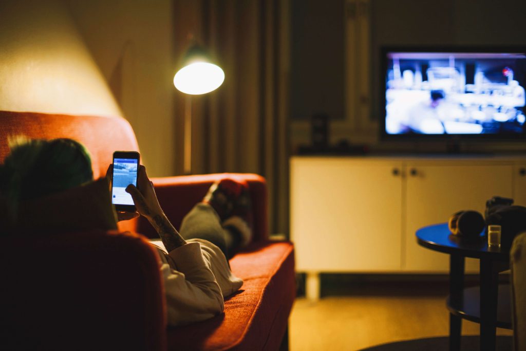 Uma mulher está deitada no sofá. Ela mexe no celular enquanto assiste TV ao mesmo tempo.