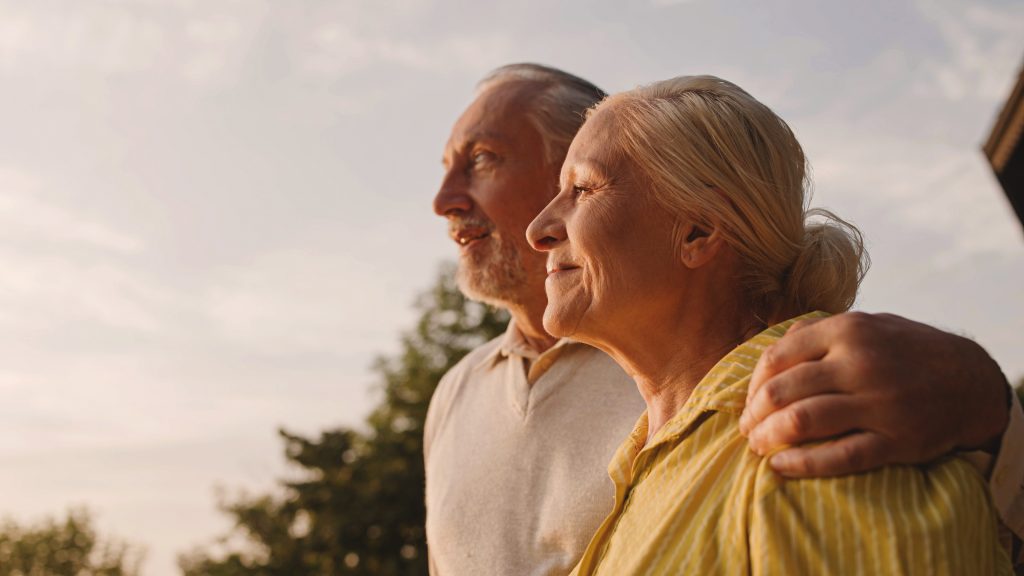 Casal de idosos sorridentes abraçando ao ar livre, curtindo vista da cidade ao pôr do sol, amor em família.