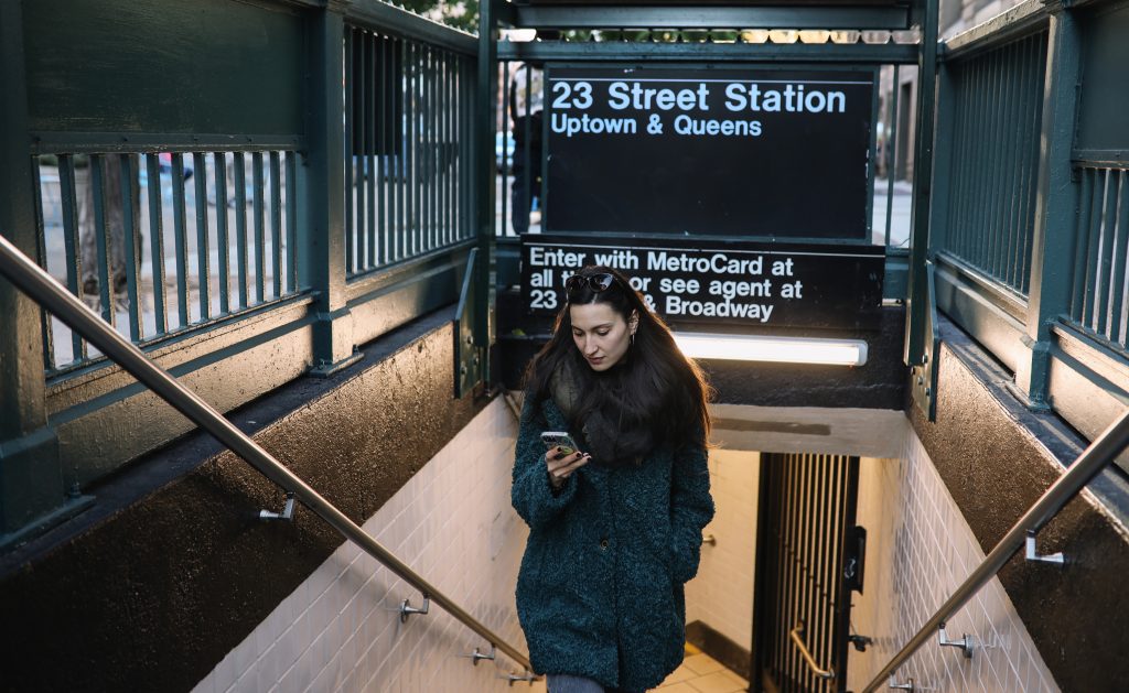 Mulher subindo o metrô de NY com um celular na mãe. Parece estar frio, pois ela está com um sobretudo e cachecol.