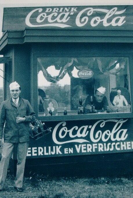 Stand da Coca-Cola distribuindo unidades da bebida de graça, nas Olipíadas de 1928. Ao lado esquerdo dele está um homem com paletó e chapeú de marinheiro, olhando para a câmera.