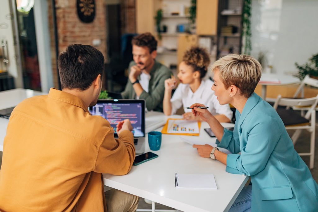 Um programador masculino mostra a uma colega uma técnica de codificação, enquanto eles montam um planejamento de mídia. Os códigos são visíveis na tela do laptop.