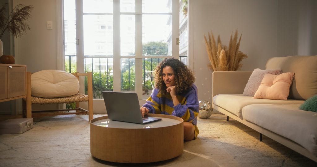 Retrato da jovem sorridente que trabalha de casa no computador portátil no apartamento ensolarado aconchegante. Empresária criativa de sucesso faz trabalho remoto para e-commerce, gerenciando pedidos on-line.