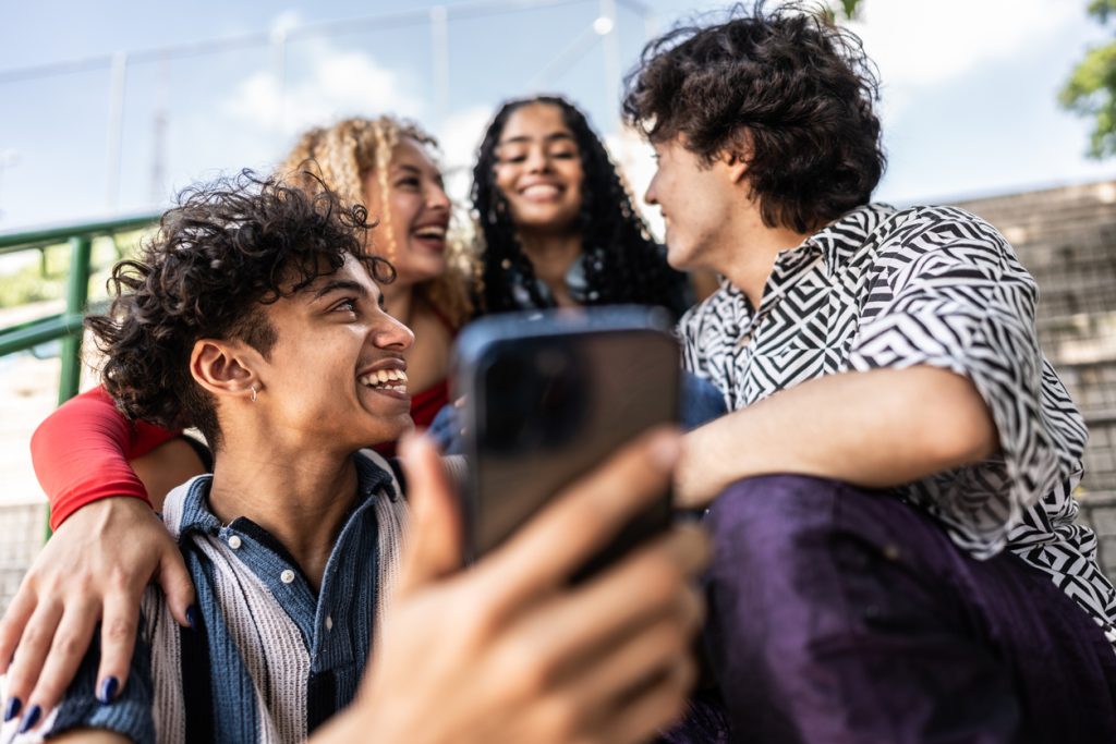 Duas mulheres estão sentadas atrás de dois homens. As quatro pessoas se olham e um deles segura um celular com a mão direita.