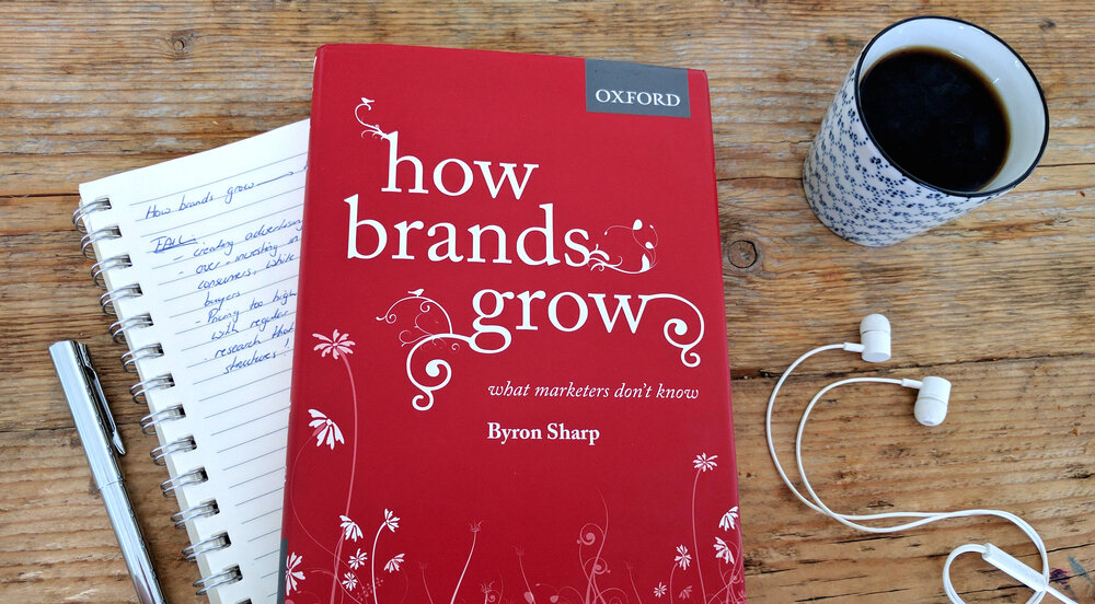 Capa do livro "How Brands Grow" e ao lado uma xícara de café e um fone de ouvido.