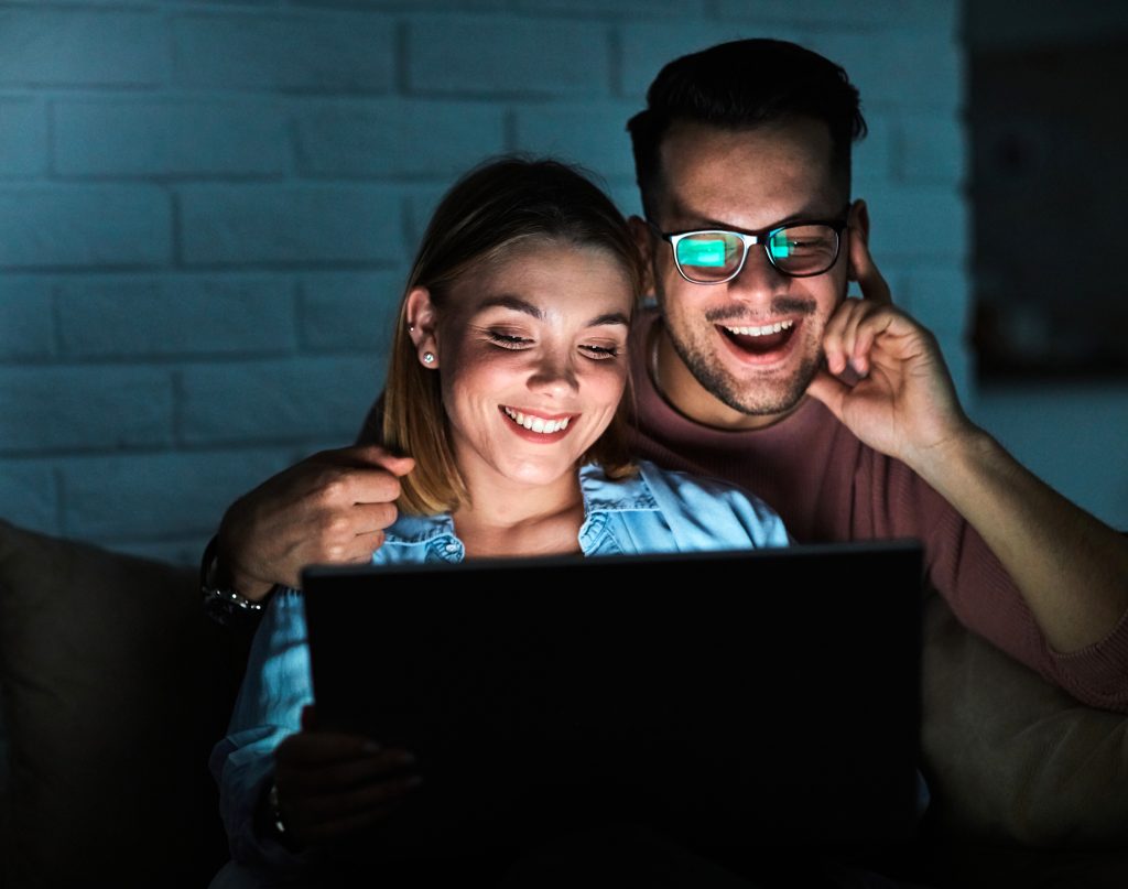 Casal jovem está sentado na frente de uma tela e sorri.