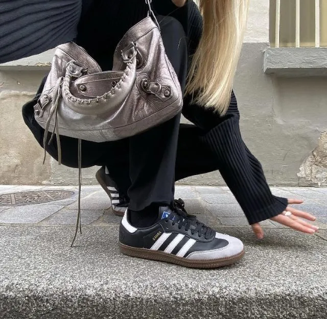 Mulher loira se abaixa para pegar algo. É possível ver sua bolsa cinza presa ao ombro. Ela usa um Adidas Samba, um ícone do marketing de nostalgia.