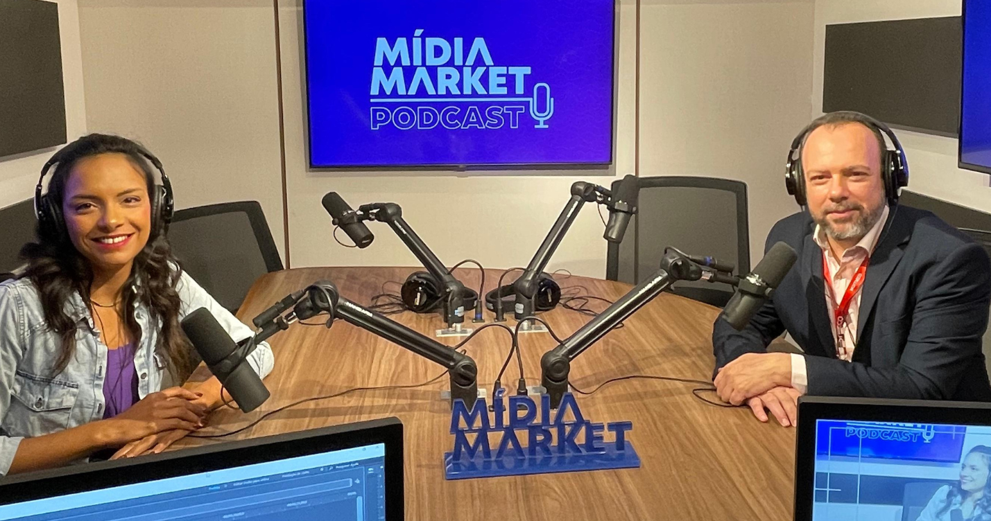 Luiza Telexa, coordenadora de mídias digitais do Grupo ND e Fernando Castellan, Gerente de Marketing da Komeco sentados em uma grande mesa de madeira de 4 lugares usando fone de ouvido posando para foto, no fundo da imagem vemos a logo do mídia market podcast.