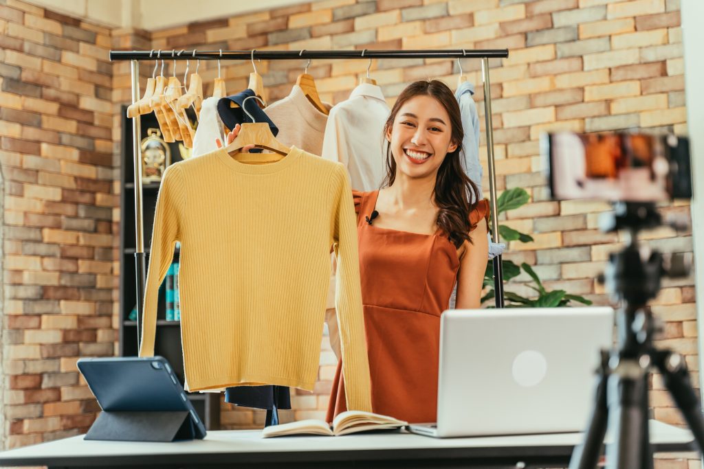 Mulher asiática segura uma blusa e aponta com ela para a tela, como quem faz um live shopping.