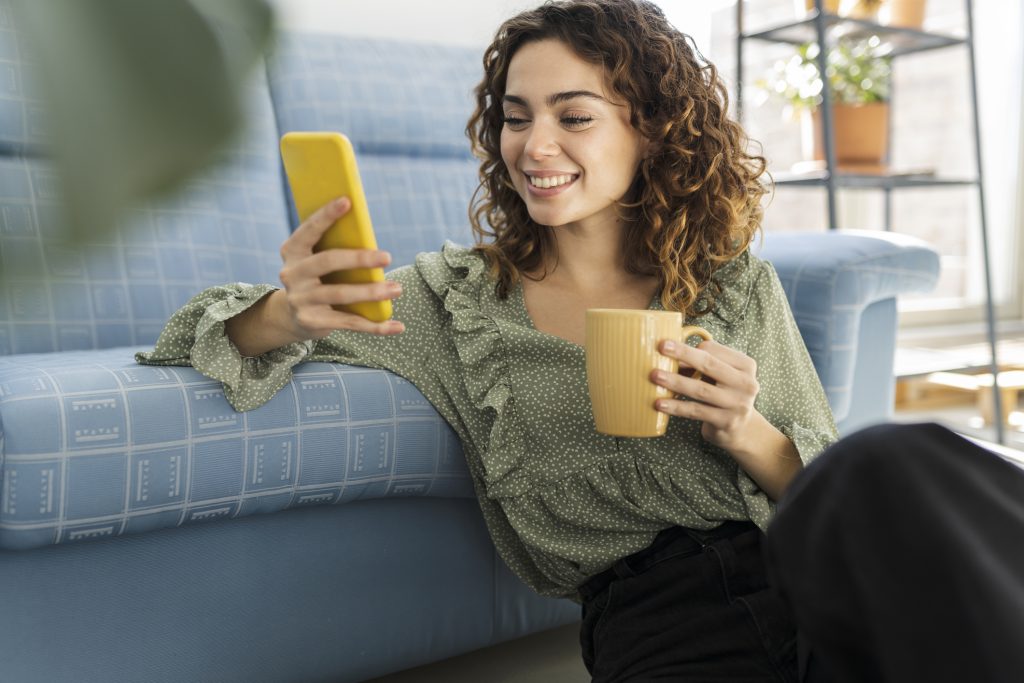 Mulher de cabelos encaracolados sentado no sofá com uma xícara de bebida quente e telefone celular.