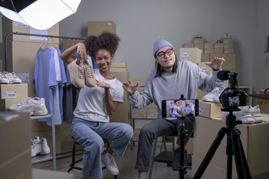 Dois jovens estão próximos um do outro e seguram um par de tênis e o mostram para a câmera. Eles estão fazendo uma live shopping.