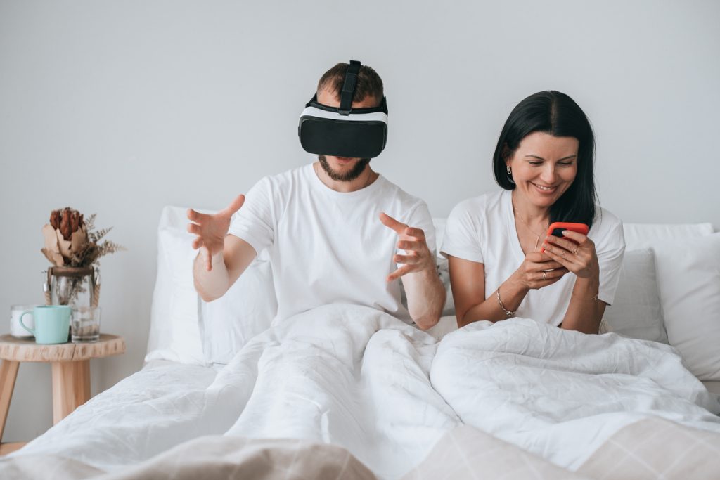 Casal na cama com homem em fone de ouvido VR gesticulando e mulher usando smartphone, divertido. A junção da realidade virtual e realidade aumentada é a realidade mista.