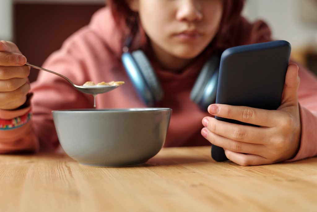 Menina cujo rosto está borrado segurando smartphone e colher de cereal.