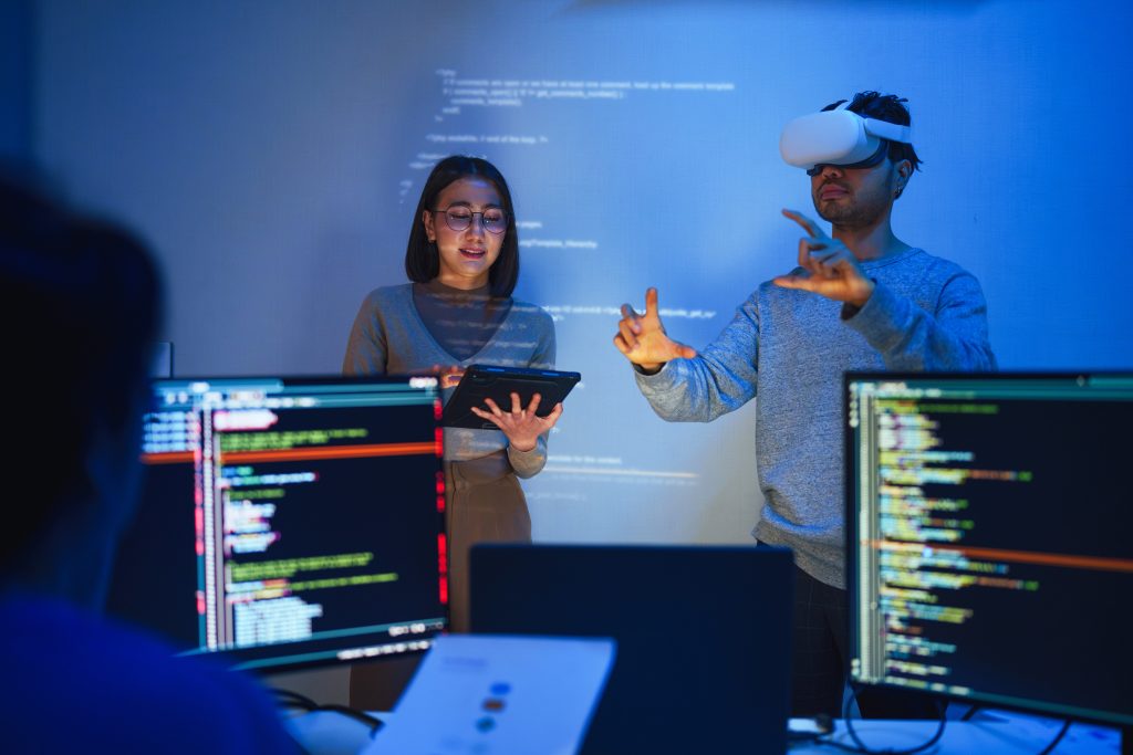 Desenvolvedor usa um headset de realidade virtual para interagir com projetos de interface projetados, enquanto colegas discutem e codificam em telas.
