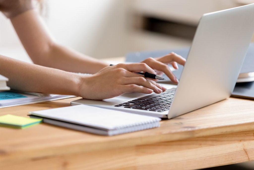 A menina está digitando no computador e só é possível ver suas mãos. Ela segura uma caneta com uma das mãos e está fazendo um briefing para influenciadores.