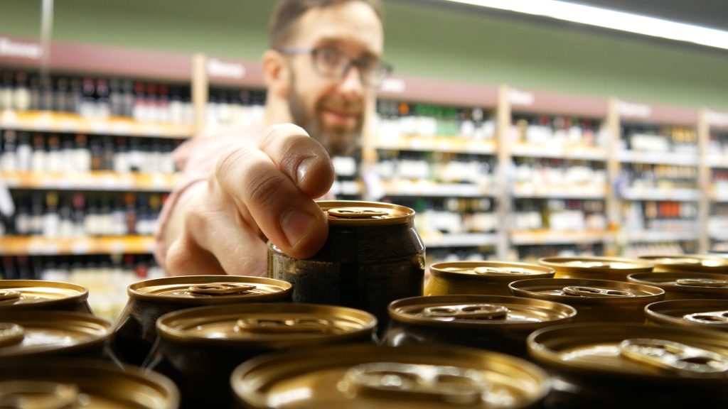 Close-up de muitas latas de cerveja em uma prateleira de loja e um homem barbudo sorridente com copos tomando um.