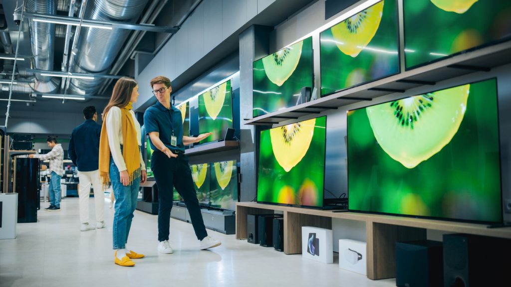 Um vendedor etá em uma loja apresentando opções de TV para uma moça. Na imagem é possível ver 6 TVs de telas grandes.