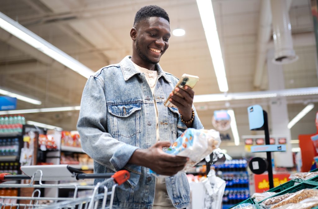 Homem negro segurando um alimento não perecível com uma mão e o celular com a outra, enquanto sorri.