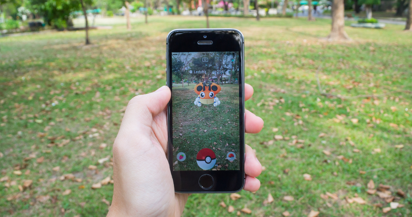 Pessoa segurando um celular no meio de um parque gramado e apontando o celular para um personagem de pokemon go que existe apenas na realidade aumentada