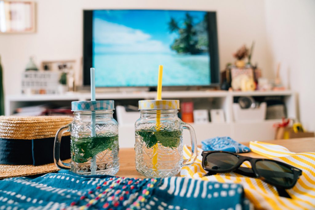 Foto conceitual de dois copos de bebida com praia paradisíaca na tela da TV em casa.