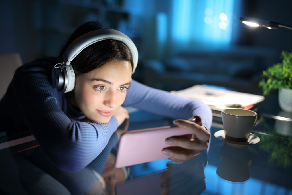 Mulher assistindo mídia no telefone inteligente à noite em casa. Ela está por dentro das tendências de streaming de 2025.