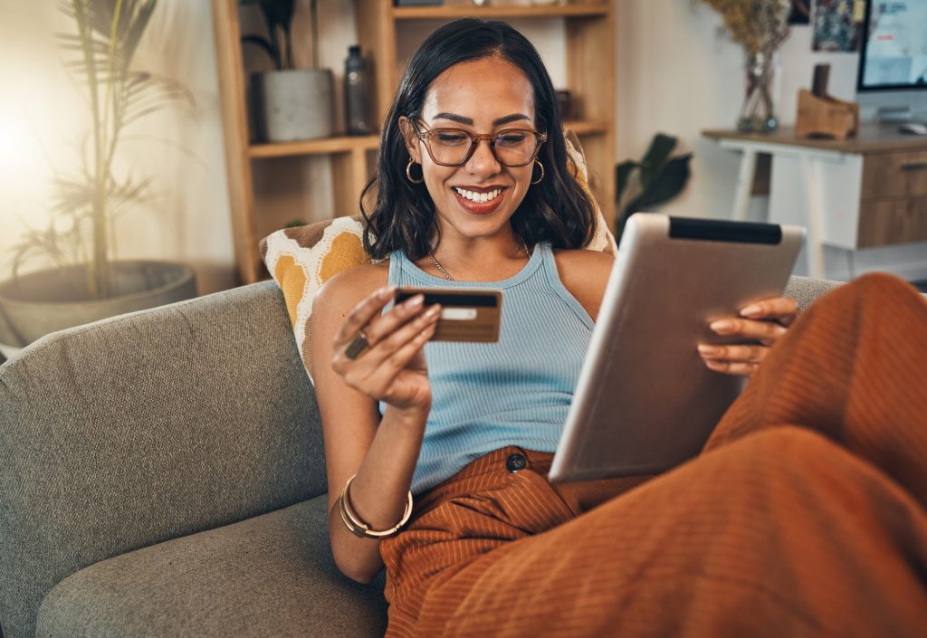 Mulher mestiça sorridente usando cartão de crédito para comércio eletrônico em tablet digital em casa.