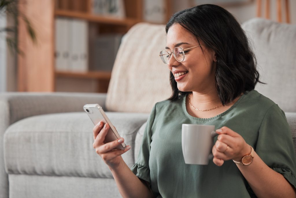 Mulher, café e digitação com telefone na sala de estar para postagem de mídia social, notificação de leitura ou bate-papo técnico.
