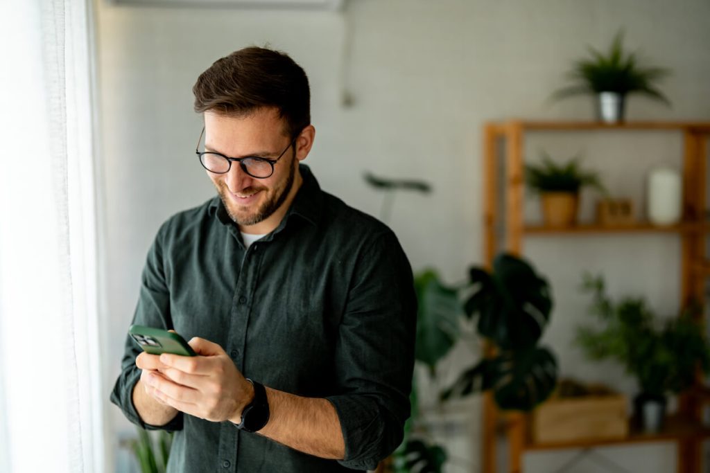 Homem branco e barbado com camisa verde segurando celular na mão.