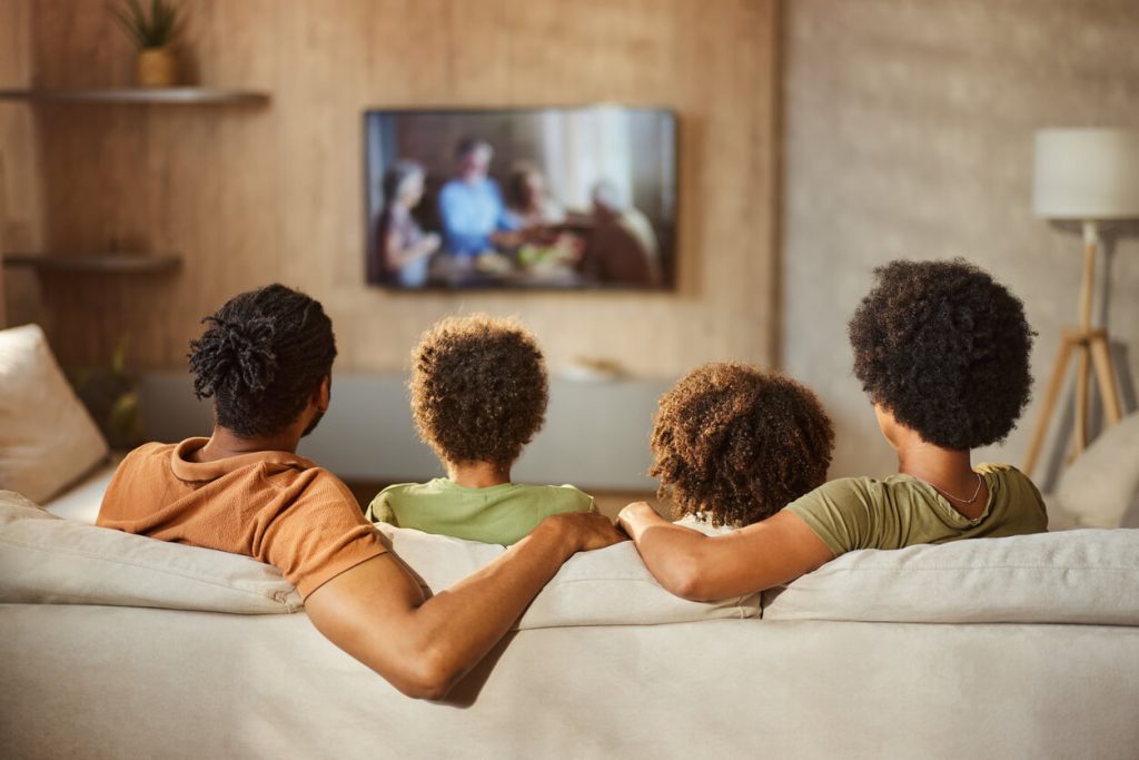 Vista traseira de uma família negra assistindo TV em casa.