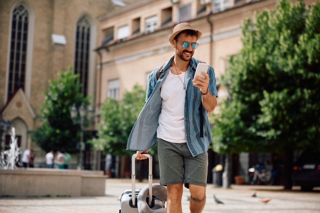 Homem feliz com saco de viagem mensagens de texto no celular enquanto caminha na cidade.