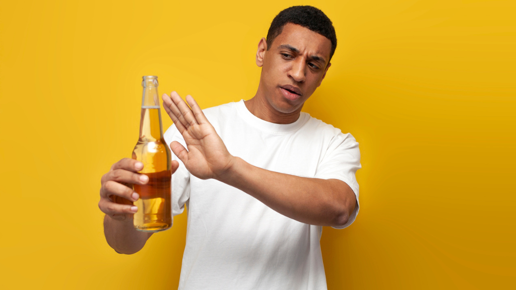 Homem jovem de camiseta branca fazendo um sinal de "pare" com a mão para uma garrafa de cerveja em um fundo amarelo, simbolizando a rejeição e a queda no consumo de álcool no Brasil.