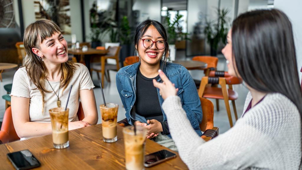 Três mulheres jovens sorrindo e conversando em uma mesa de café/bar moderno, consumindo bebidas geladas sem álcool, ilustrando a mudança de hábito e a redução do consumo de álcool no Brasil.