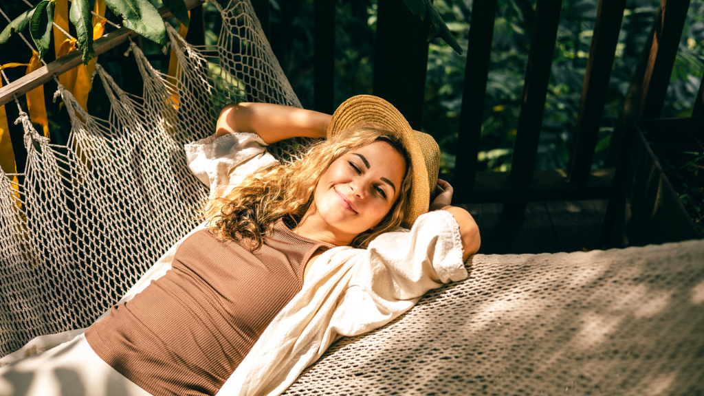 Uma mulher jovem e sorridente relaxa em uma rede de descanso sob a luz do sol, usando um chapéu de palha. A imagem ilustra o comportamento do consumidor que busca repor energias, um dado essencial para estratégias de marketing de carnaval voltadas ao bem-estar.